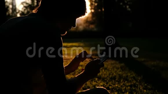日落时分一个手里拿着手机的男人的剪影他正在草地上用一包纸打一条短信视频的预览图
