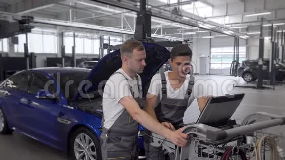 成熟的机械师和他的助手在汽车修理站用电脑视频的预览图