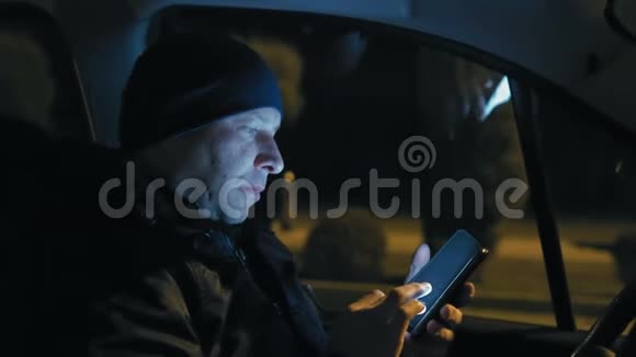 特写镜头男人在停车的夜晚坐在汽车的车轮前看智能手机上的新闻视频的预览图