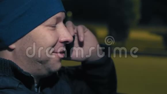 特写镜头男人在电话上说话微笑在停车场的夜晚坐在车轮旁视频的预览图