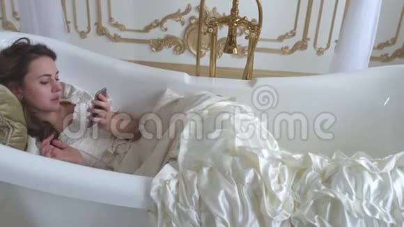 可爱的年轻女人躺在柔软的枕头上的空浴室里手里拿着羽毛扇子用手机说话视频的预览图