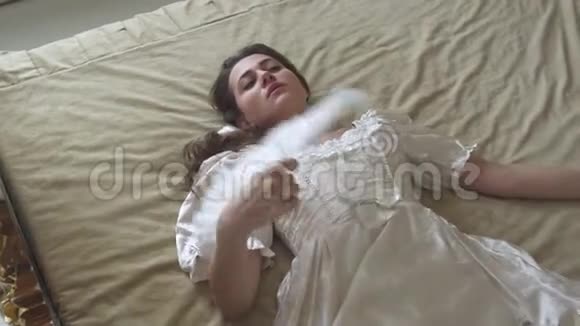 年轻漂亮的女人穿着白色的老式球裙躺在巨大的金色床上用羽毛扇挥动着视频的预览图