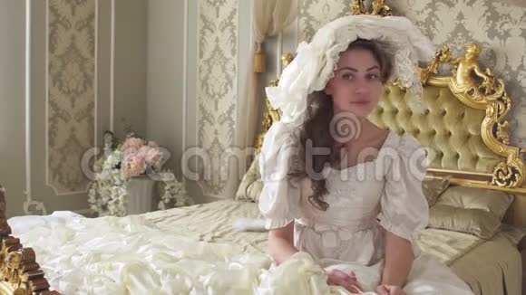 年轻漂亮的女人穿着白色的老式球裙和帽子坐在巨大的金色床上用手机躲起来视频的预览图