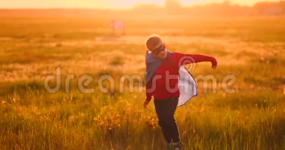 一个穿西装的男孩和一个超级英雄面具在草地上日落时跑过田野视频的预览图