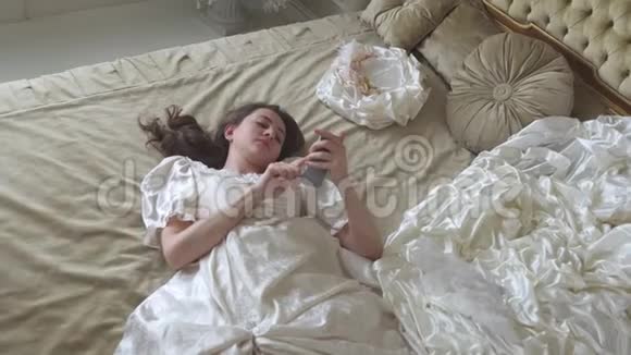 年轻疲惫的女人穿着球袍躺在金色装饰的床上在手机上发短信视频的预览图