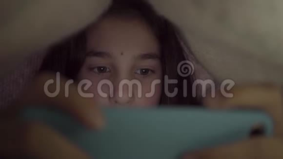 可爱的少女玩她的手机躺在毯子下关闭睡觉时间蓝光有负面影响视频的预览图