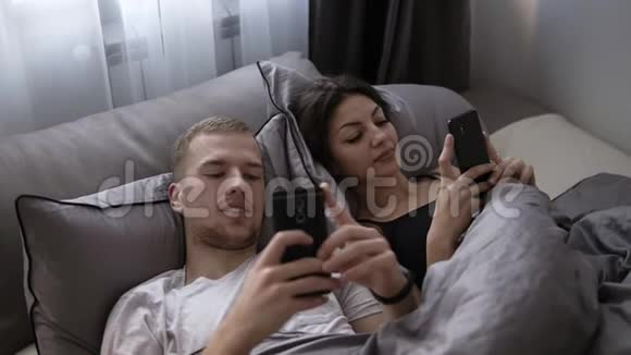 年轻的已婚夫妇在床上躺着互相微笑时使用手机快乐的生活方式情侣床视频的预览图