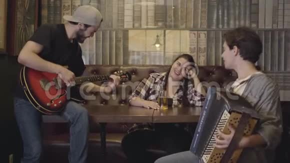 年轻的笑着留着胡须的男人在酒吧里弹吉他他的朋友在手风琴上演奏而漂亮丰满的女人坐着视频的预览图