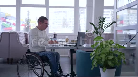 电子商务成功的成熟男性轮椅上的残疾人戴眼镜使用笔记本电脑坐在咖啡馆的桌子上视频的预览图