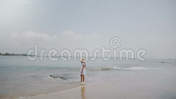 大气背景拍摄的快乐小女孩在异国情调的海洋海滩上漫步戴着大草帽视频的预览图