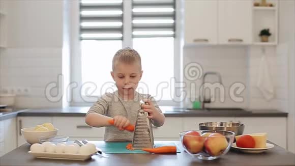 漂亮的白种人小男孩大蓝眼睛在明亮的厨房里做饭他是一根胡萝卜视频的预览图