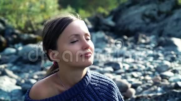 阳光明媚的日子里穿着毛衣的漂亮女人坐在河边的石头上把脸闭上女孩眼睛视频的预览图