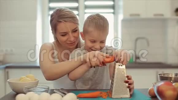 妈妈教儿子搓胡萝卜他们在笑一位年轻漂亮的母亲穿着白色衬衫可爱的儿子穿着白色衬衫做饭视频的预览图