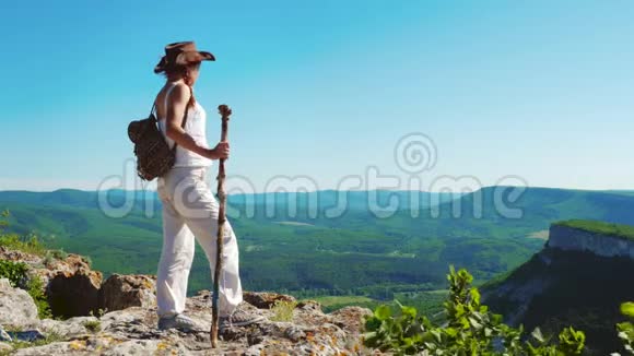 穿着牛仔帽和白色衣服的女旅行者站在山顶上欣赏着令人惊叹的景色视频的预览图