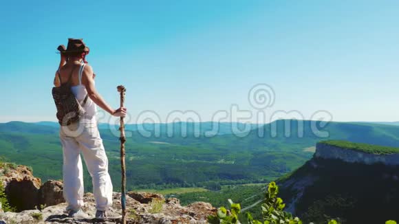 穿着牛仔帽和白色衣服的女旅行者站在山顶上欣赏着令人惊叹的景色视频的预览图