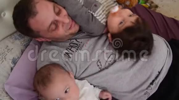 父亲和两个儿子婴儿和蹒跚学步的孩子一起在床上玩耍视频的预览图