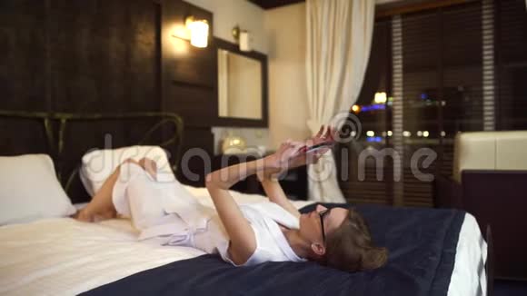 漂亮的女商人戴着眼镜躺在床上手里拿着平板电脑视频的预览图