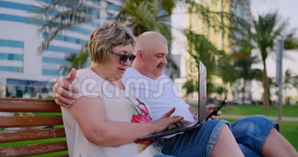 今年夏天在一个现代化的城市里快乐的老夫妇坐在长凳上手提电脑放在摩天大楼的背景上视频的预览图
