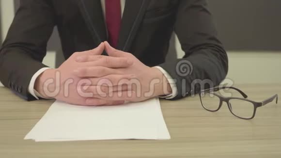 一个穿着西装手指交叉的人坐在桌子前手里拿着白纸钢笔和视频的预览图