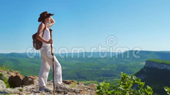 穿着牛仔帽和白色衣服的女旅行者站在山顶上欣赏着令人惊叹的景色视频的预览图