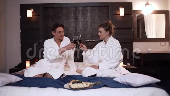 漂亮的一对穿着浴袍喝酒的夫妇坐在现代酒店房间的床上家庭休闲视频的预览图