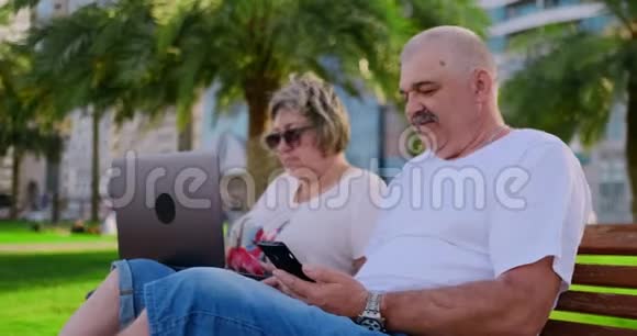 今年夏天在一个现代化的城市里快乐的老夫妇坐在长凳上手提电脑放在摩天大楼的背景上视频的预览图