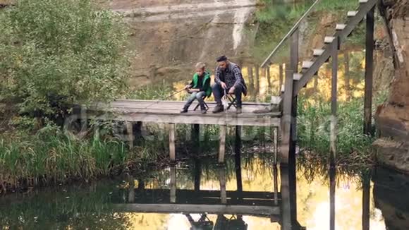 穿着休闲服装的帅哥和他的孩子坐在木船坞上用木棒钓鱼视频的预览图
