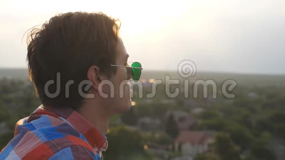 戴墨镜的英俊男子站在屋顶欣赏傍晚美丽的景色年轻人休息视频的预览图