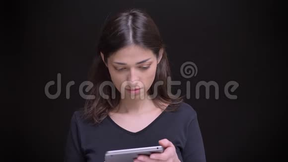 白种人长发黑发女孩的肖像专注地注视着平板电脑的微笑进入黑色背景的相机视频的预览图