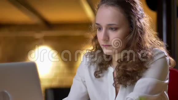 一位年轻的卷发女士的特写肖像她专心地用笔记本电脑工作并在办公室里把笔记录在笔记本上视频的预览图