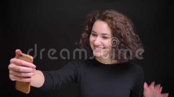 一位年轻迷人的白种人女性的特写照片她在电话中打着视频微笑着挥手问好并在打电话视频的预览图