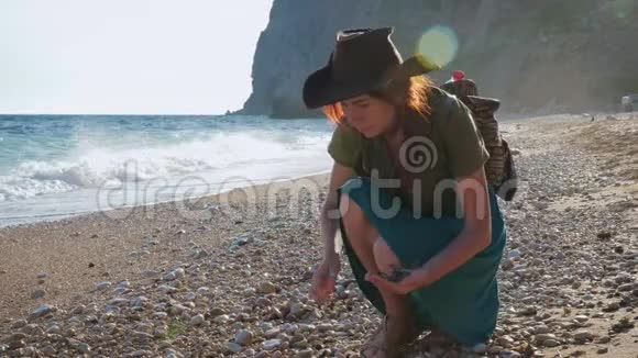年轻的红发女孩旅行者带着牛仔帽和背包漫步在海边视频的预览图