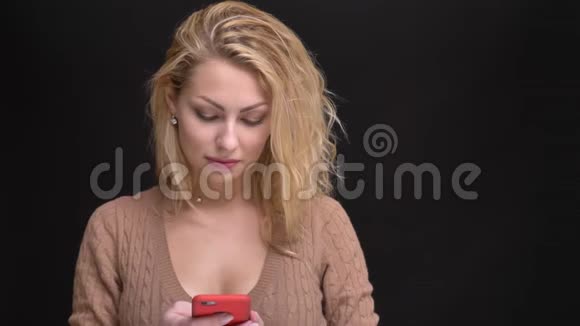 美丽的白种人长发女子的肖像微笑着观看黑色背景下的智能手机视频的预览图