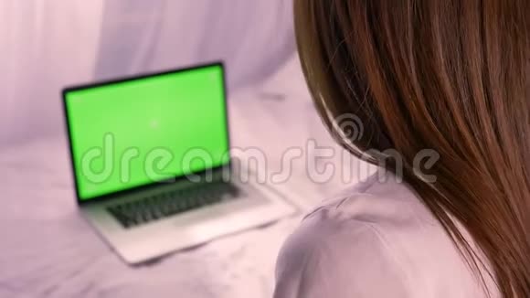 年轻的深色头发的女性在家中坐在床上观看笔记本电脑的绿色彩色按键屏幕视频的预览图