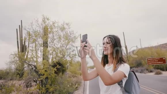 在国家公园远足期间慢动作年轻快乐微笑的旅行者女性在仙人掌沙漠上拍摄智能手机照片视频的预览图