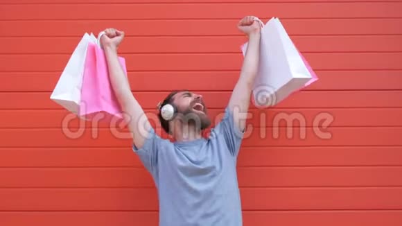 一个积极时髦的胡须男人的肖像他拿着粉红色和白色的购物袋在红色的背景上听音乐的人视频的预览图
