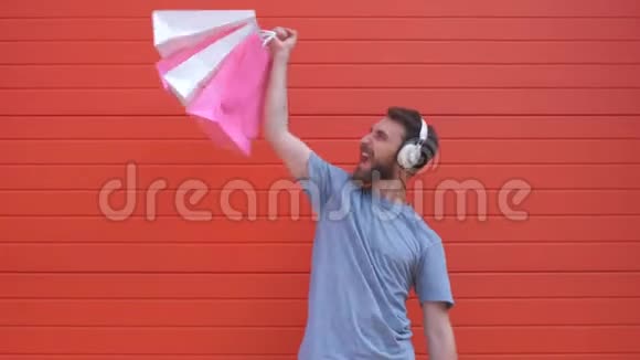 一个积极时髦的胡须男人的肖像他拿着粉红色和白色的购物袋在红色的背景上听音乐的人视频的预览图