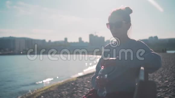 一个穿着运动衫和眼镜的黑发女孩坐在靠近大海的长凳上从塑料瓶里喝水视频的预览图