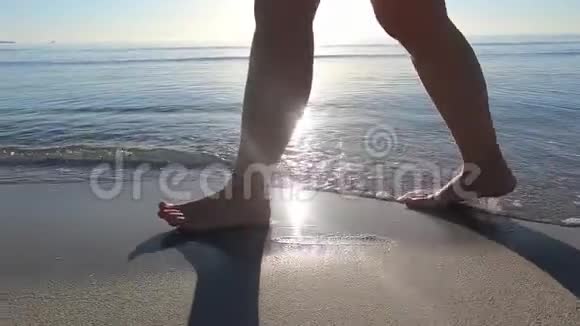 一个年轻女人的腿沿着海岸走着海浪拍打着她的脚在黎明时的背光下踩着沙子视频的预览图