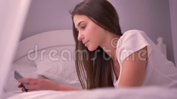 年轻漂亮的少女用平板电脑发送信息躺在床上微笑的特写镜头视频的预览图