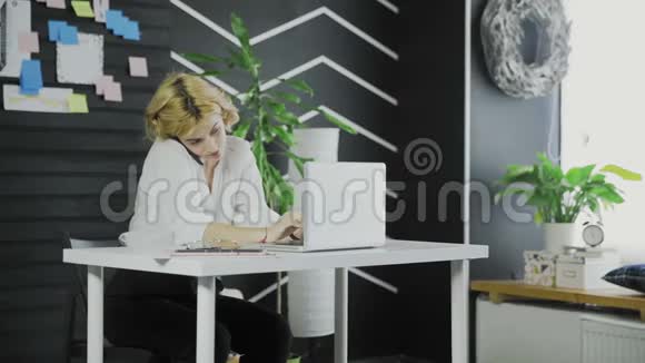 漂亮的商务女性在办公室里用手提电脑键盘打字时在手机上说话视频的预览图