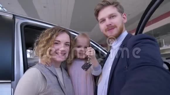 家庭自拍照片在汽车商店有趣的夫妇与可爱的小女孩与钥匙拍照在智能手机附近新购视频的预览图