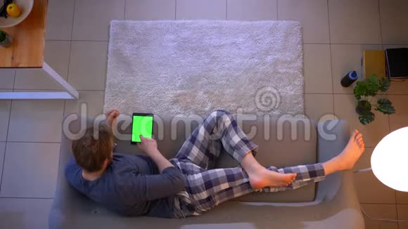 年轻的穿着随意的男性在室内躺在沙发上用绿色屏幕的平板电脑特写镜头视频的预览图