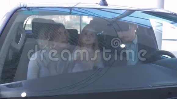汽车经销店的幸福家庭肖像年轻父母带着小女孩微笑拥抱坐在车里视频的预览图