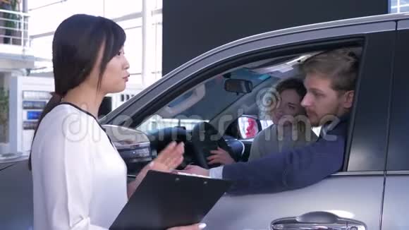 卖车的亚裔女性在展厅买车时会咨询坐在汽车沙龙中的年轻家庭消费者视频的预览图