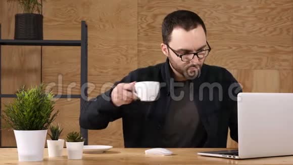 在笔记本电脑上工作时喝咖啡或茶的留胡子的年轻人视频的预览图