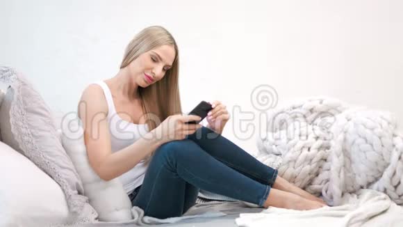 坐在舒适的沙发上用智能手机写信息的自信忧郁的年轻美女视频的预览图