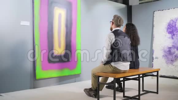 男人和女人坐在长凳上在画廊里看抽象的图画视频的预览图