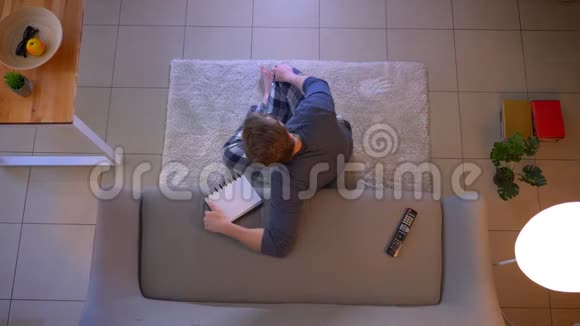 一位穿着随意的年轻男士坐在地板上看电视在室内写下食谱的特写镜头视频的预览图