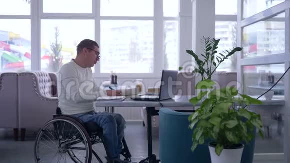 电子学习坐在轮椅上的老年男性残疾人戴着眼镜阅读书籍并使用笔记本电脑坐在咖啡馆的桌子上视频的预览图
